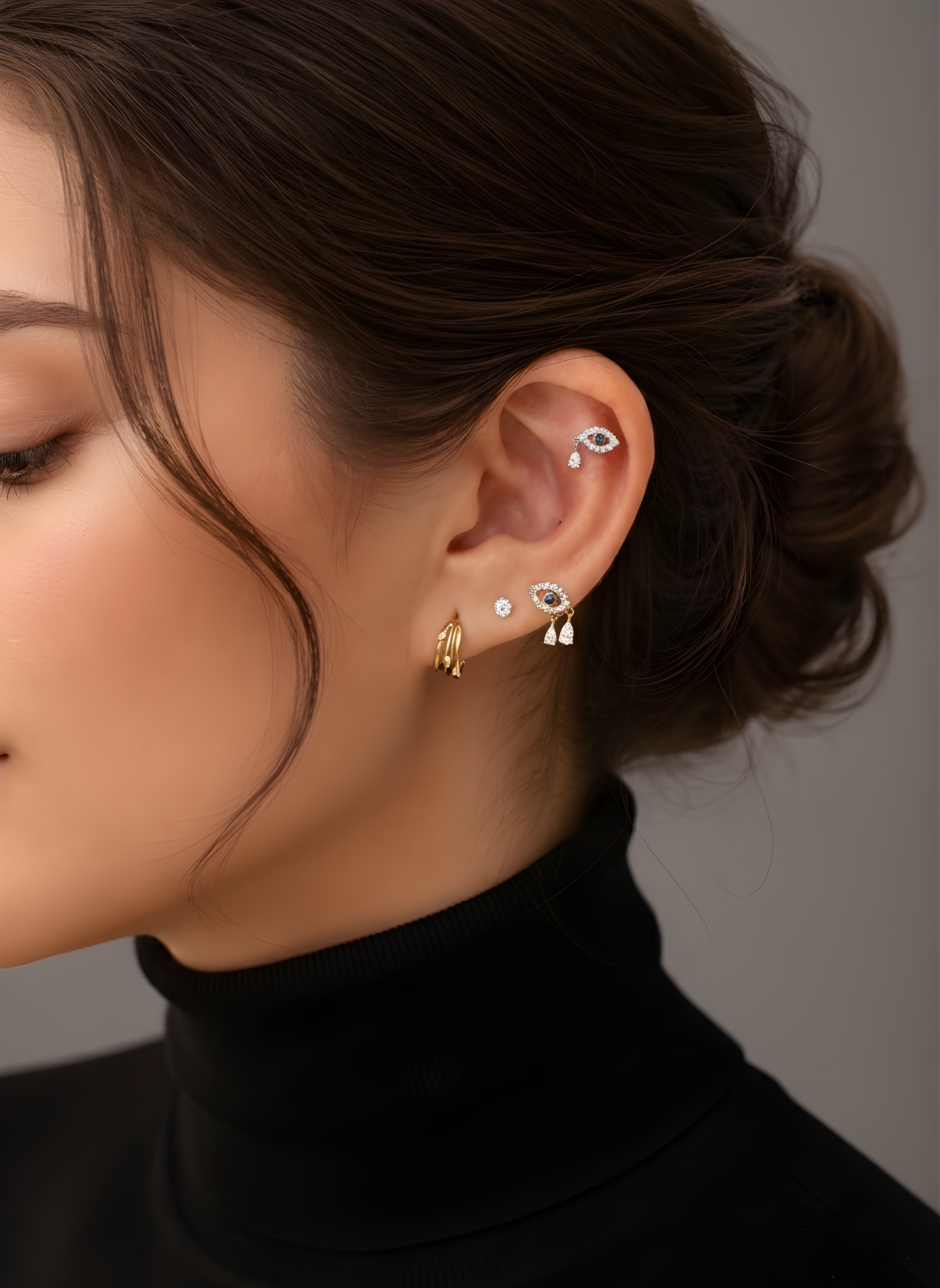 A woman's ear with a curated stack featuring Lark & Berry's gold Evil Eye earrings with diamond drops and a helix stud.