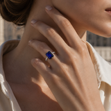 A woman's hand with the London Bridge Blue Sapphire Ring, featuring an emerald-cut sapphire and a pavé diamond band, on her neck.