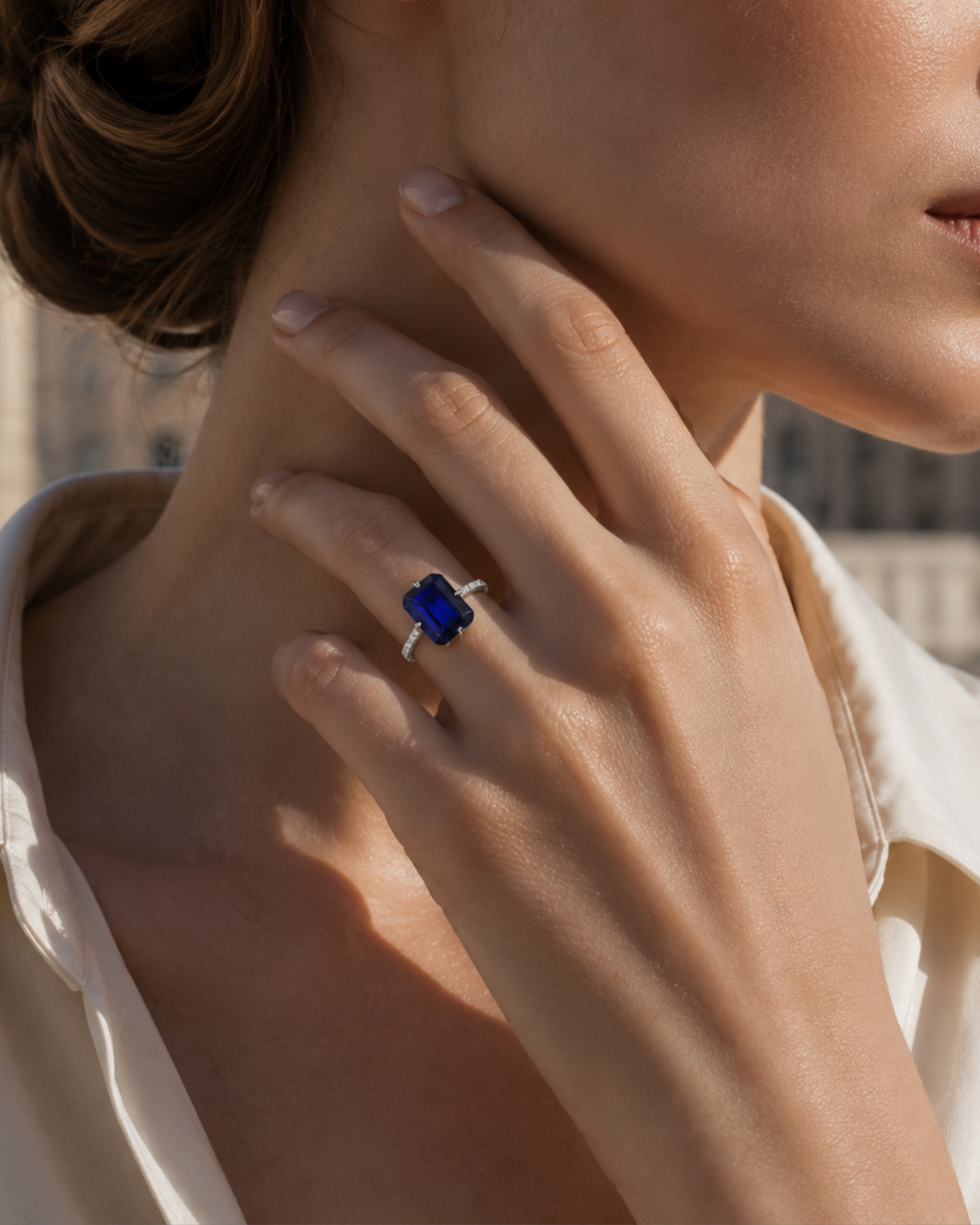A woman's hand with the London Bridge Blue Sapphire Ring, featuring an emerald-cut sapphire and a pavé diamond band, on her neck.